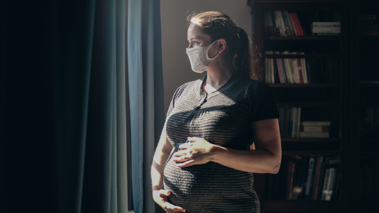 Pregnant woman with face mask standing in front of window.