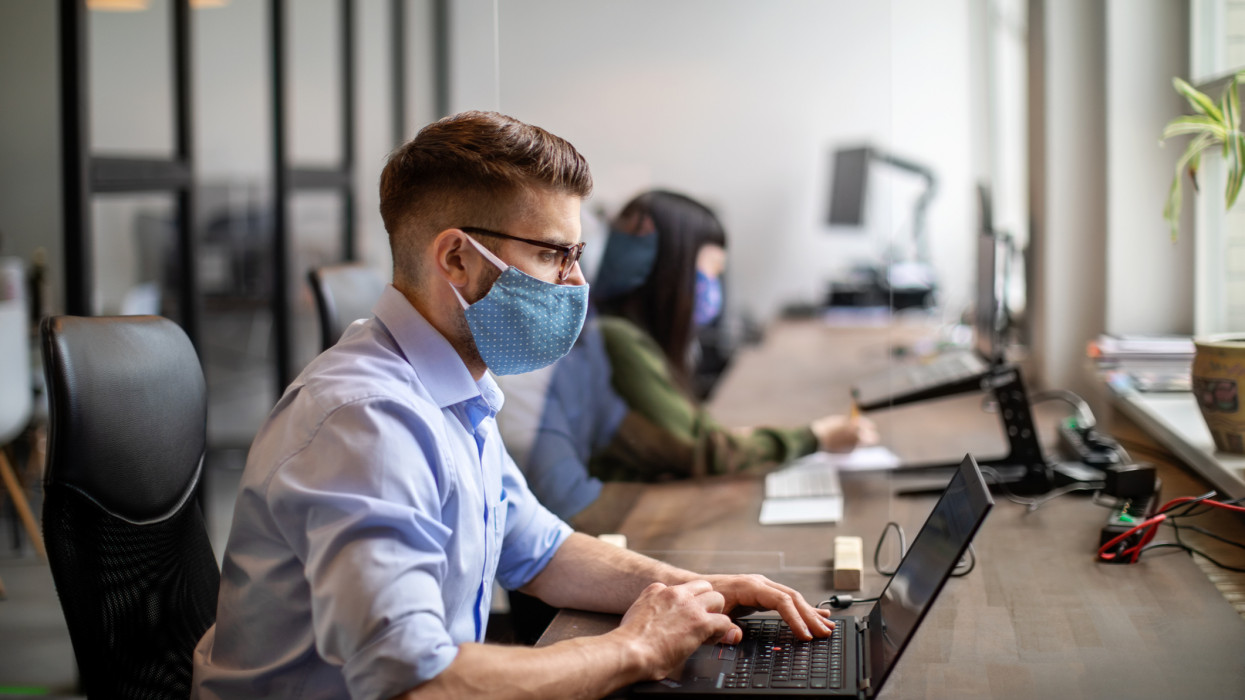 Businessman wearing protective face mask working at his desk. Business people back to work after pandemic sitting at desk with protection guard between them.