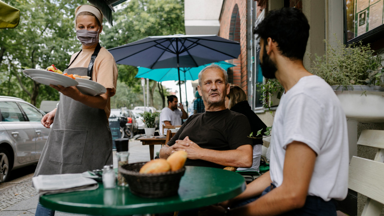 A waitress wearing a face mask serving some food to customers sitting outdoors at a restaurant.