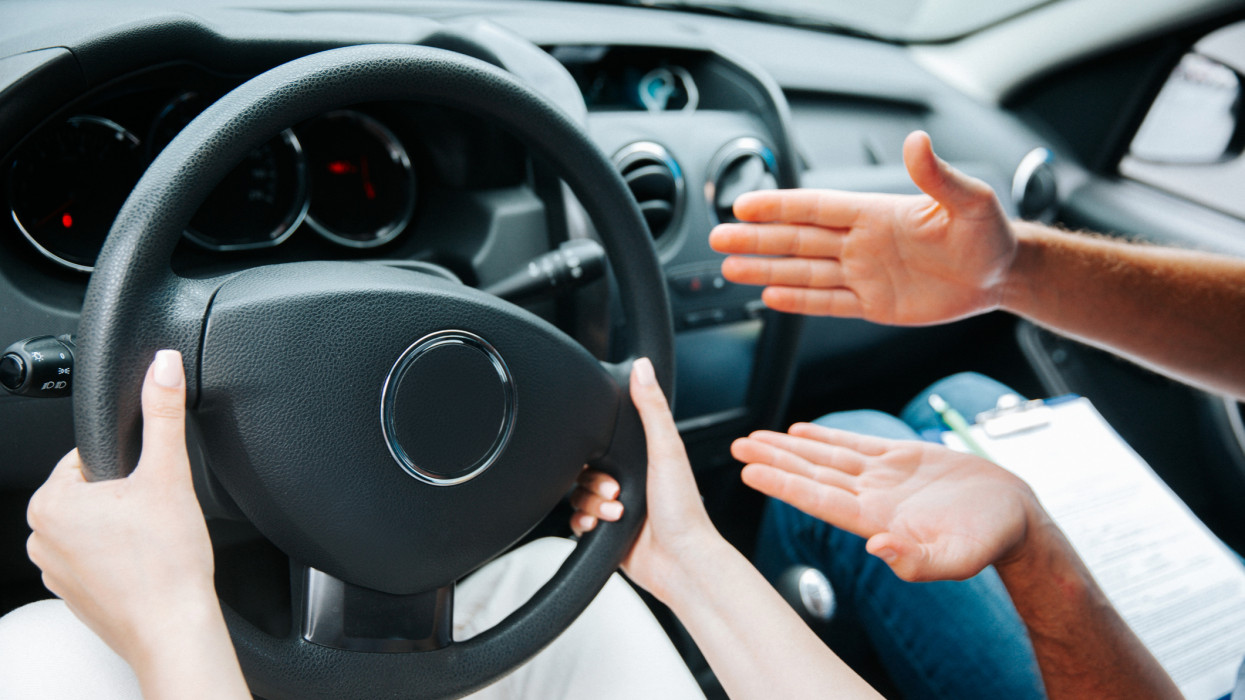 Female hands hold steering wheel. Male instructor demonstrates with his hand what to do with a wheel. Cut close-up view of woman driving a car. Driving courses concept