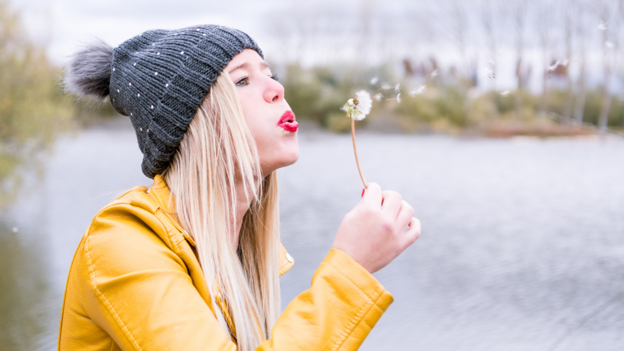 Blonde woman with red lips and grey wool cap blowing a dandelion to the right, small particles come out of the dandelion. Background with water and trees without autumn leaves.