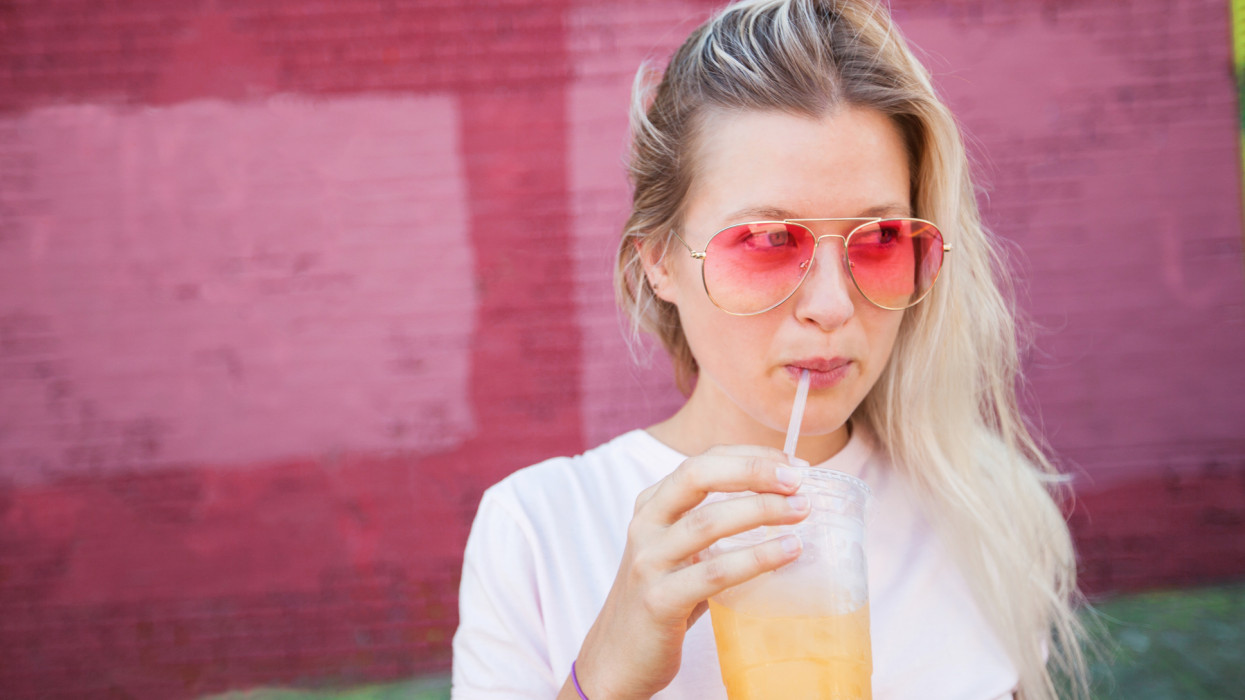 Young blonde woman wearing pink sunglasses drinking juice from a a plastic cup and straw in front of a pink wall