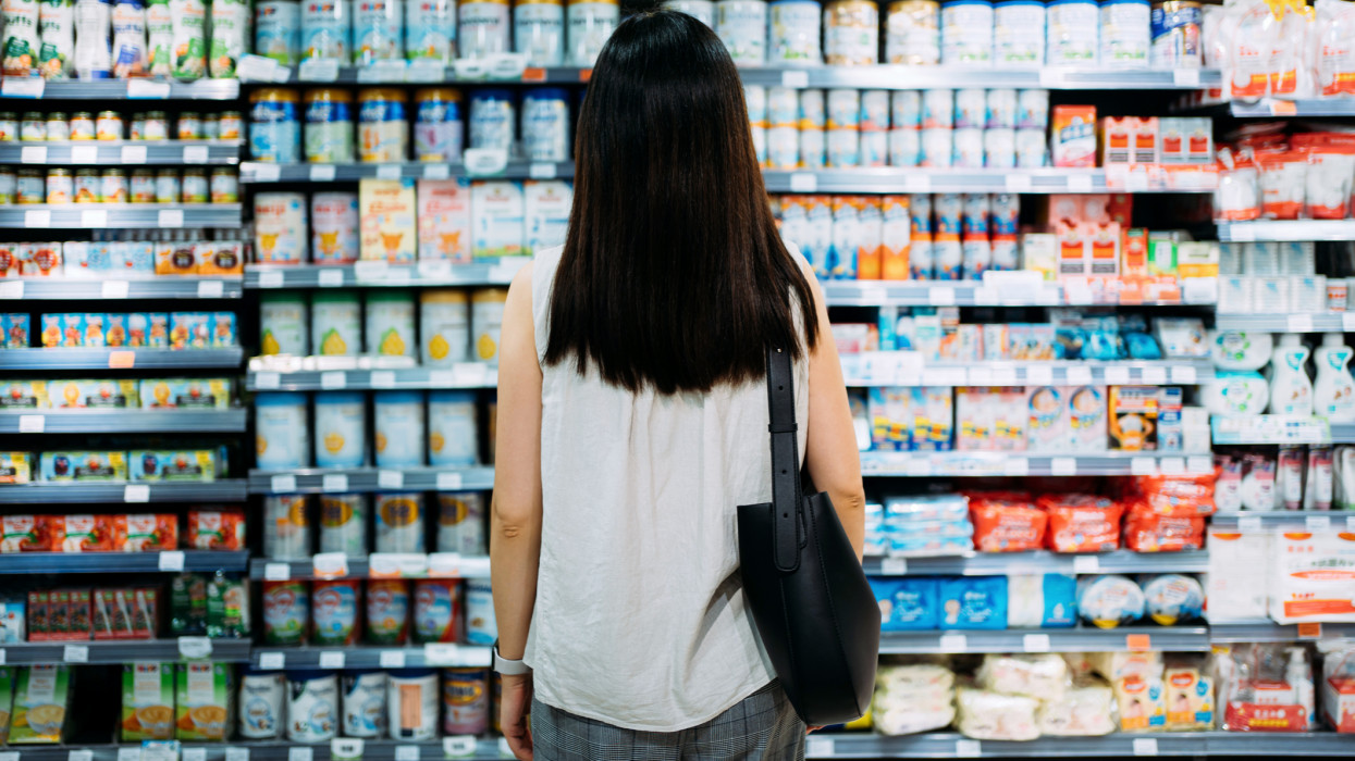 Rear view of young Asian mother groceries shopping for baby products in a supermarket. She is standing in front of the baby product aisle and have no idea which product to choose from