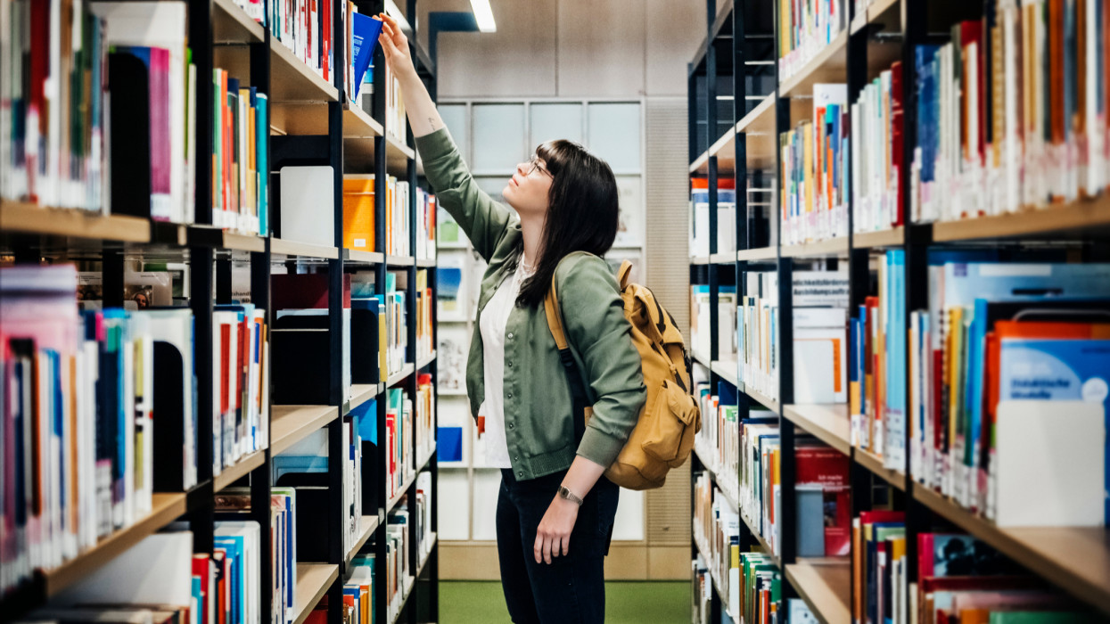 A young student reaching to select a book from the top shelf in a public library.