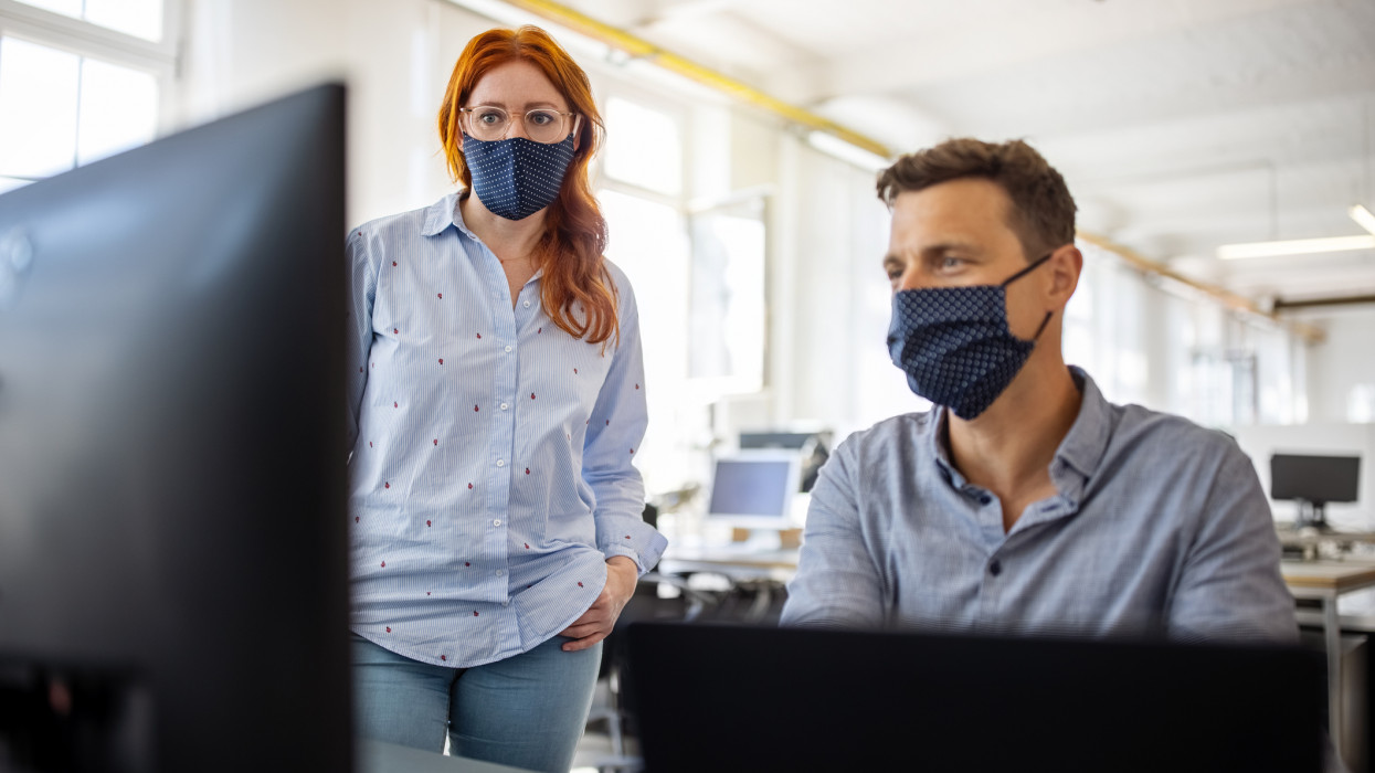 Businessman working on a computer with a female colleague standing by in the office. Business colleagues wearing face masks working together on a computer.