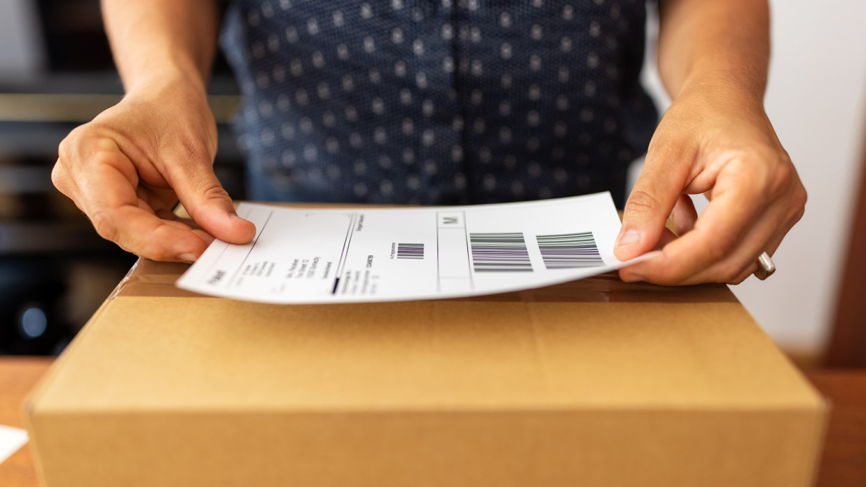 Close-up of a female hands sticking a label on a box for delivery . Woman online business owner preparing package for shipping at home.