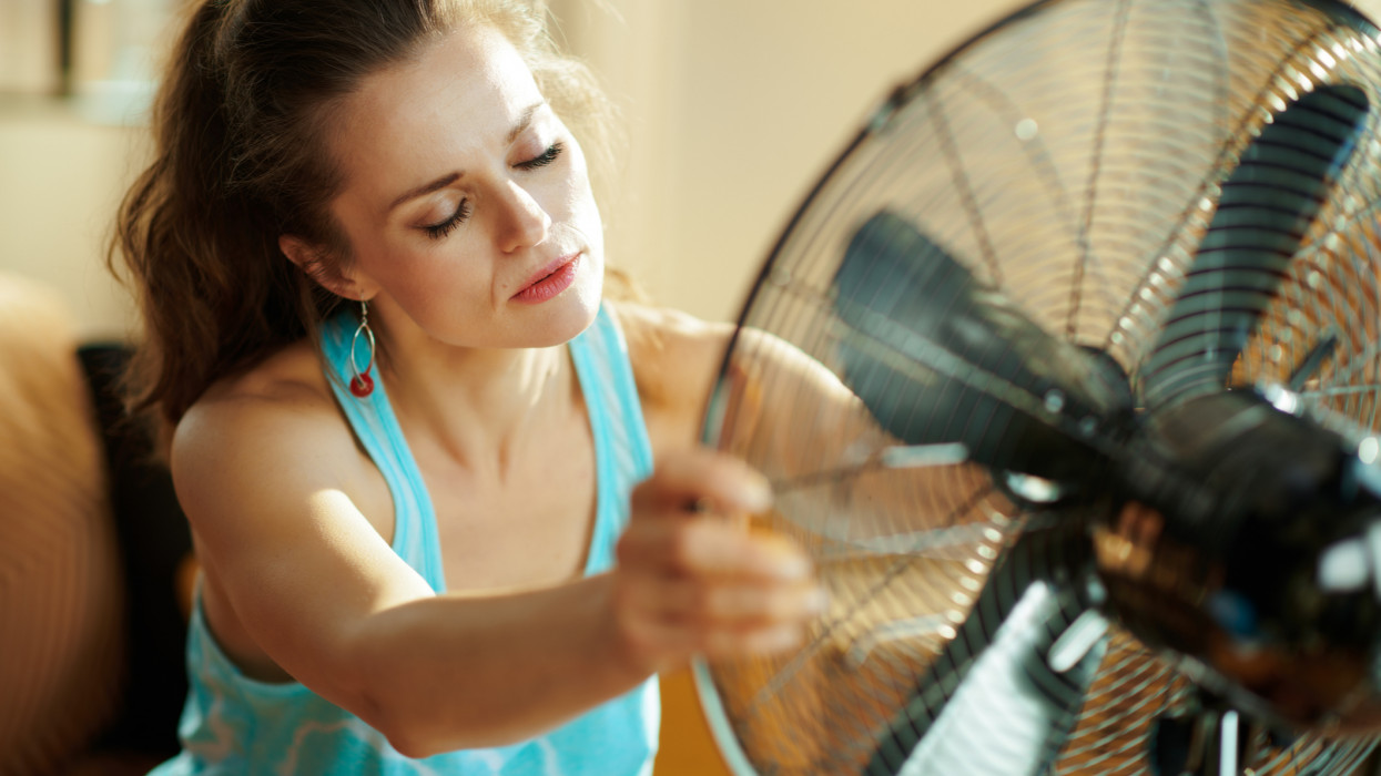 unhappy young housewife in the modern living room in sunny hot summer day enjoying freshness in the front of fan suffering from summer heat.