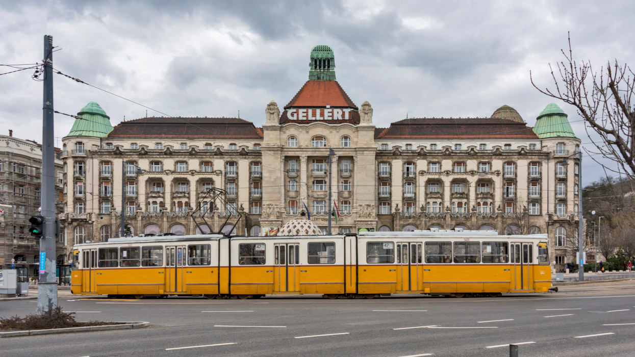 A yellow tram, and commuters in front of the Historical building of part of the famous GellÃ©rt baths in Buda, the GellÃ©rt Thermal Baths and Swimming Pool is a bath complex in Budapest, Hungary.
