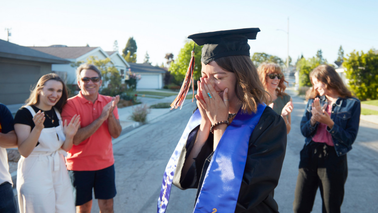 A 22 year old caucasian young woman dressed in a graduation hat and gown is overcome by emotion while surrounded by her friends and family in front of her home during celebrating her graduation from college