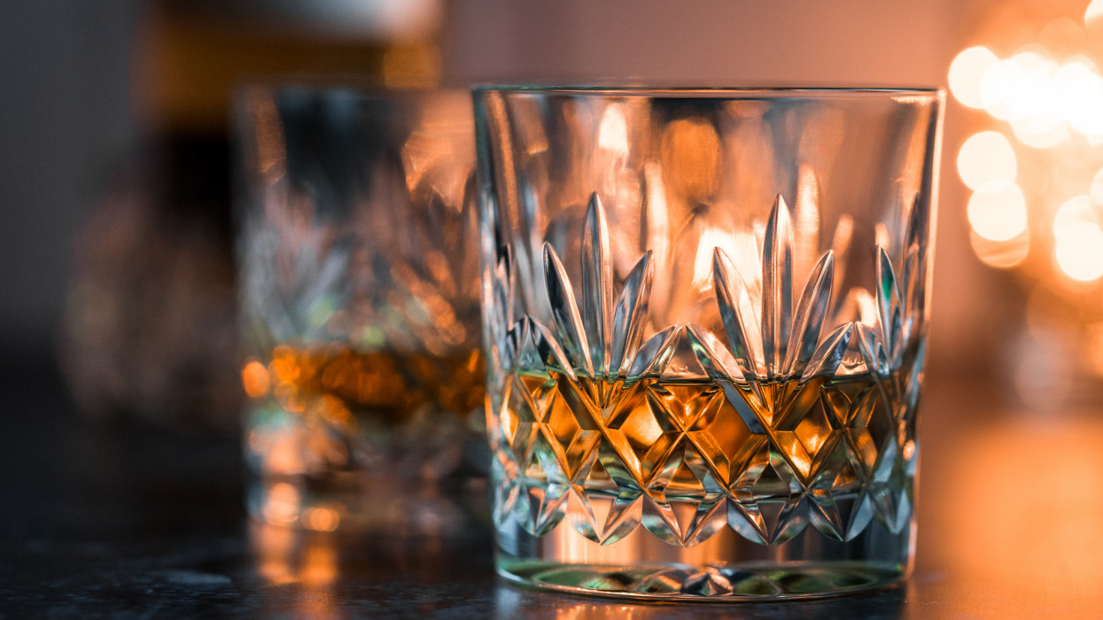 Close up color image depicting two fine crystal glasses of malt whisky on a white wooden surface. The glasses of whisky are surrounded by whisky paraphernalia such as a glass decanter and a hip flask. Room for copy space.