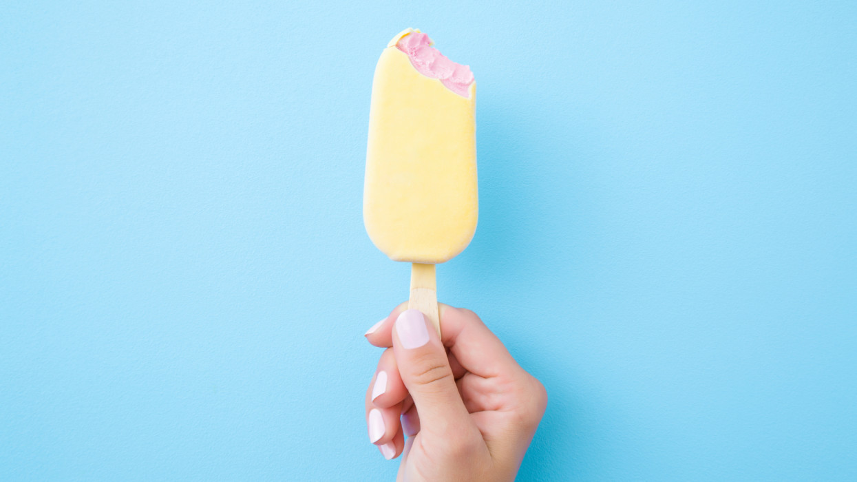 Young woman hand holding pink ice cream with white chocolate glaze on pastel blue background. Bitten food. Closeup. Top view.