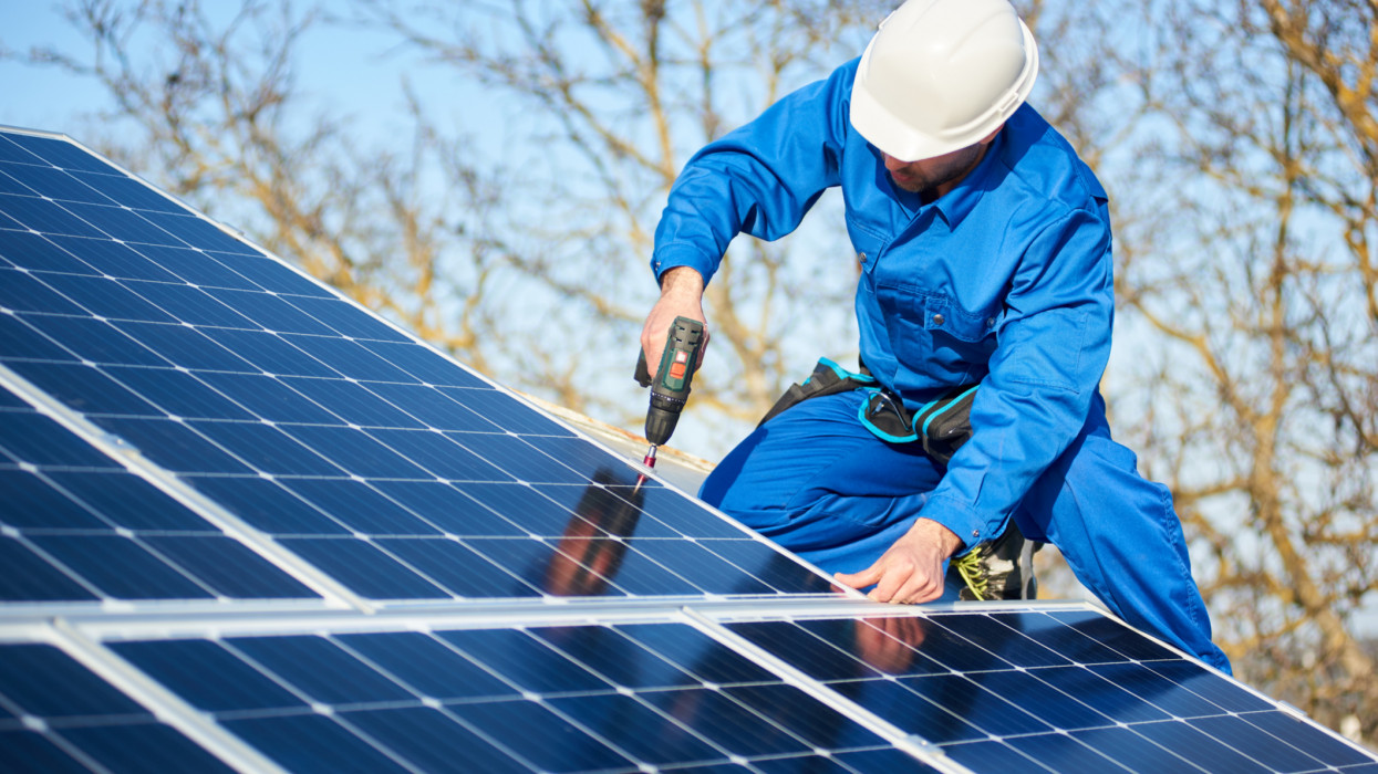 Male engineer in blue suit and protective helmet installing photovoltaic panel system using screwdriver. Professional electrician mounting solar module on roof. Alternative energy ecological concept.