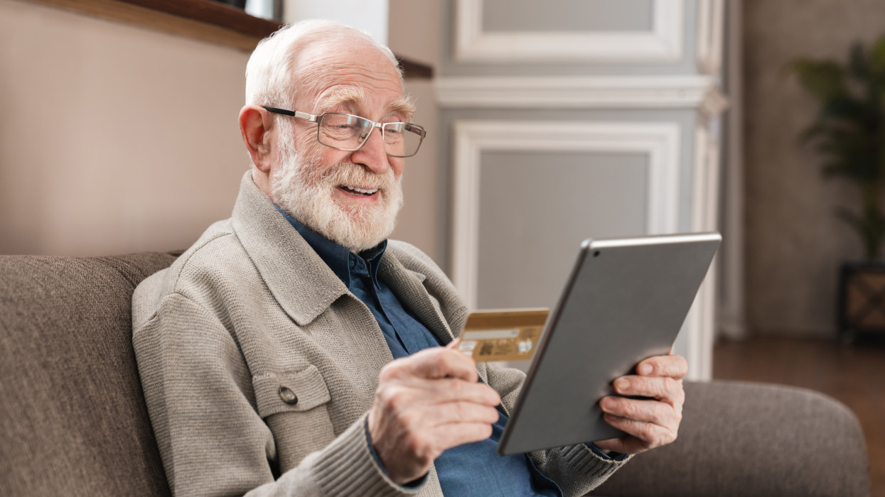 Cheerful old grandfather man doing online payment from digital tablet with credit card. Elderly senior caucasian grandpa paying bills online with credit card