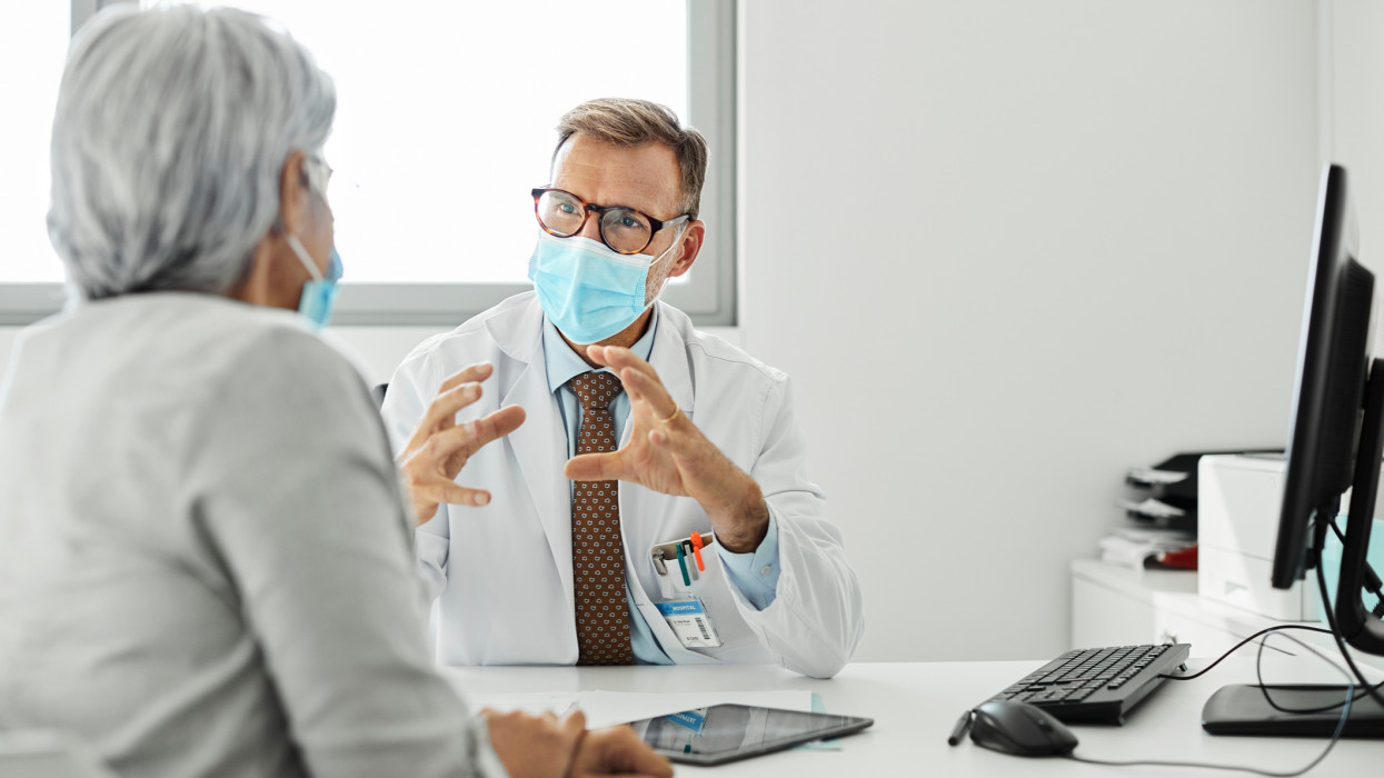 Doctor discussing with female patient during COVID-19. Male medical professional is explaining woman while sitting at desk. They are wearing protective face masks in clinic.