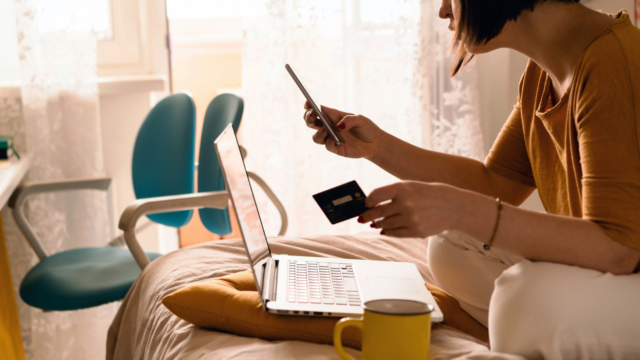 A middle-aged woman in white jeans and a yellow sweater sitting on the bed in a yoga pose in front of a laptop and a cup of coffee. Remote work at home. Online shopping in quarantine period cimlapi