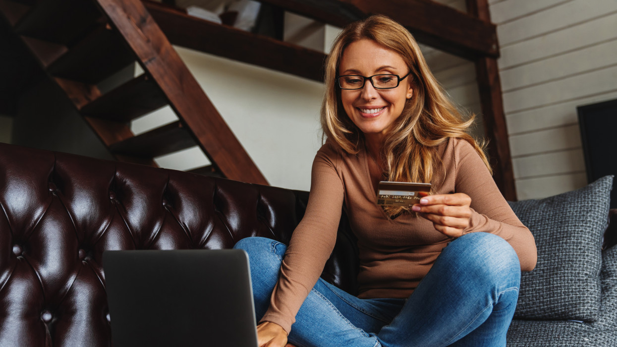Smiling woman with credit card and laptop sitting on sofa at home. E-commerce concept
