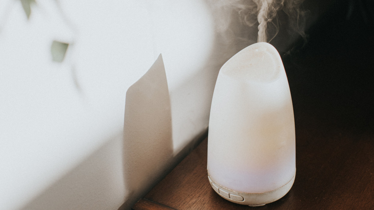 A simple white oil diffuser spritzing a light mist of water and aromatherapy blended oils into the air. It creates a shadow on the wall. Space for copy.