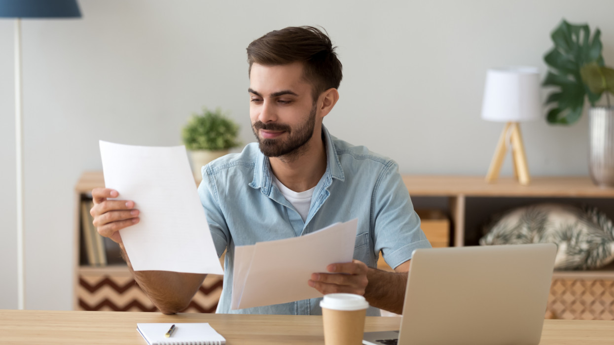 Satisfied man reading paper documents, letters, receiving pleasant news, working with laptop at workplace, sitting on desk, successful businessman, freelancer looking at contract with good offer