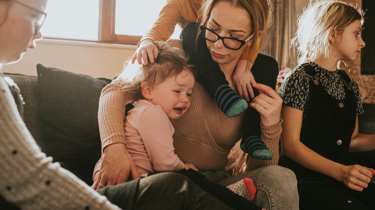 A single mother with four children sits on a sofa with her kids. Her little boy sits on her shoulders as her toddler snuggles into her as she cries. Mum consoles her. The scene is stressful, as the parent attempts to multitask. A recognisable scene that parents can relate to.