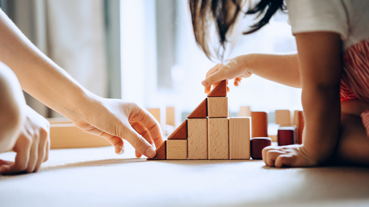 Close up of mother and little daughter sitting on the floor playing with wooden building blocks together and enjoying family bonding time at home