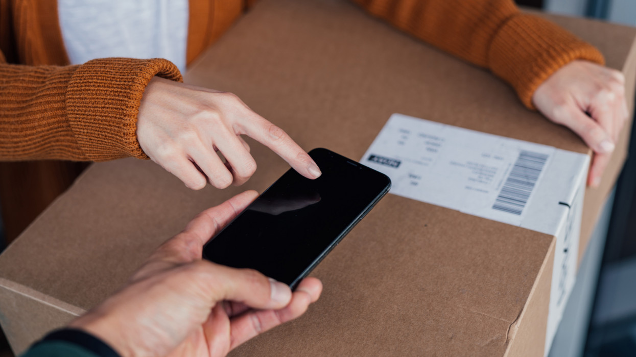 Close-up of young woman holding a carton box, signing for her delivery on a digital device from the courier. Home delivery service makes life easier. Online shopping concept.