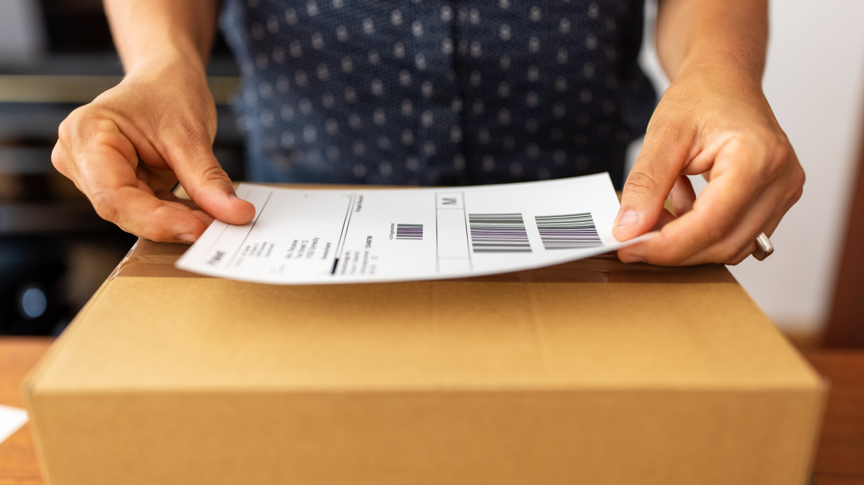 Close-up of a female hands sticking a label on a box for delivery . Woman online business owner preparing package for shipping at home.