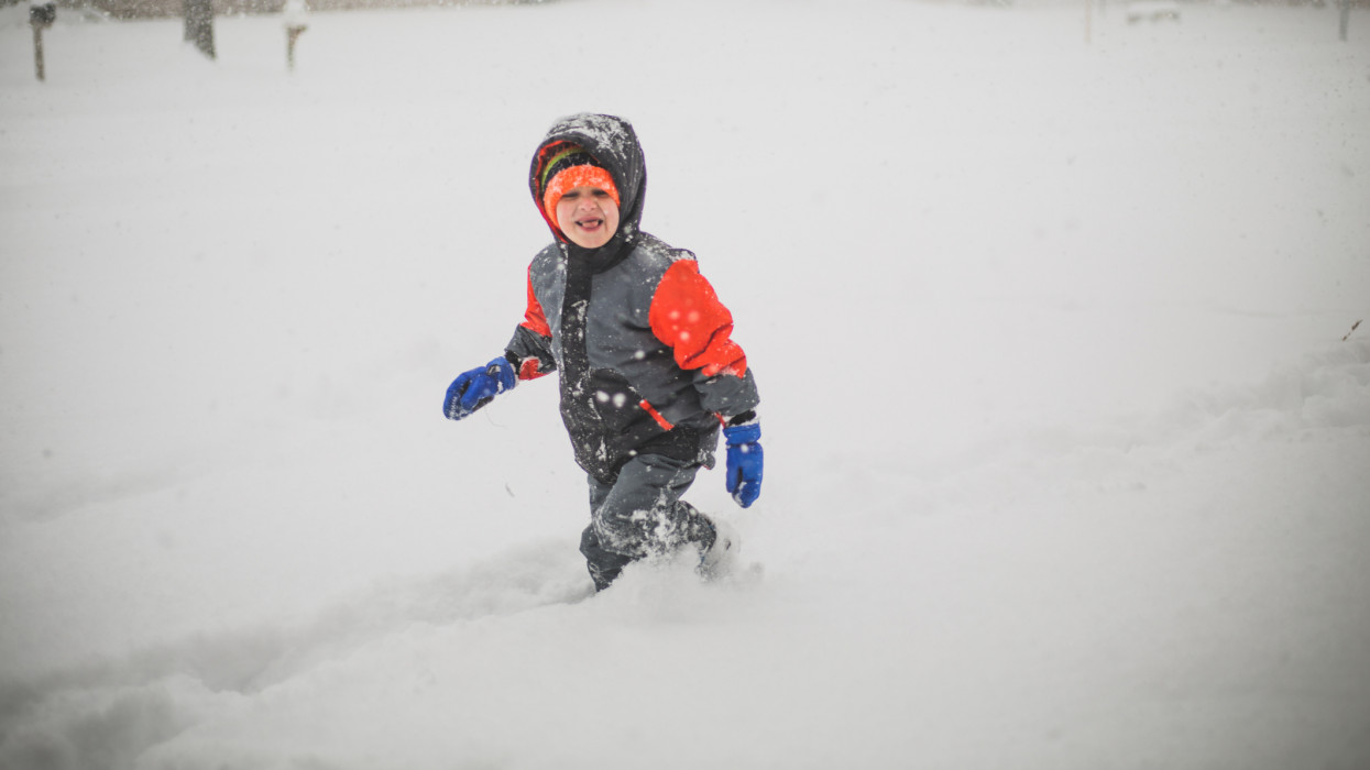 Little 3 year old boy stands outside bundled up during a snow storm and makes silly happy faces at the camera.
