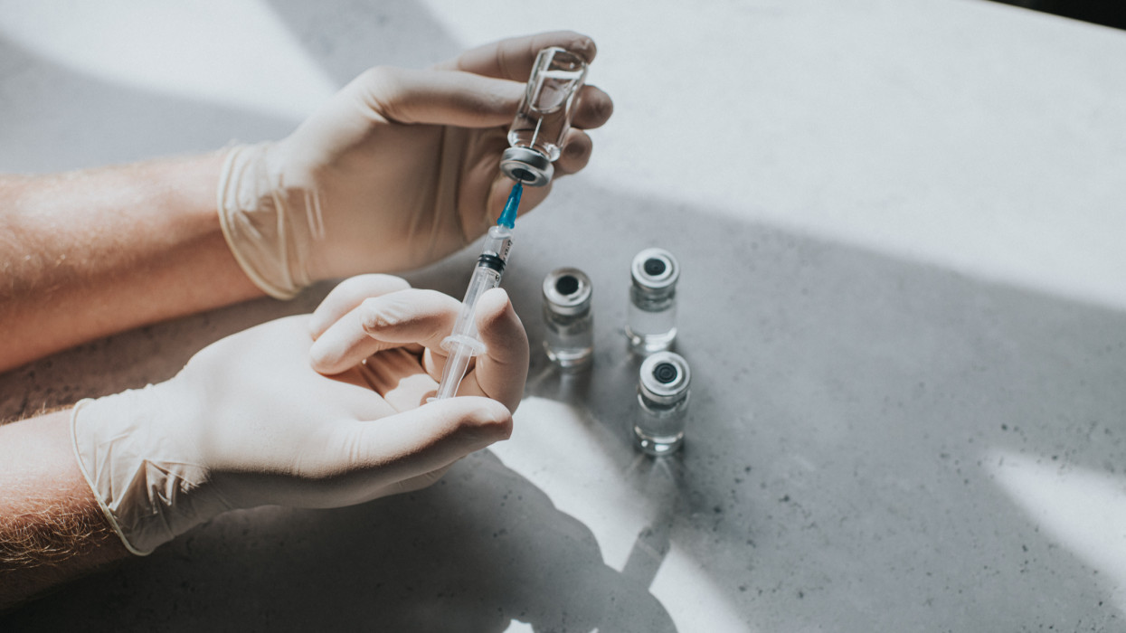 A hand wearing latex gloves hold a phial of clear liquid and a syringe. The medical professional uses the syringe to extract the medicine from the glass bottle. Conceptual with space for copy.