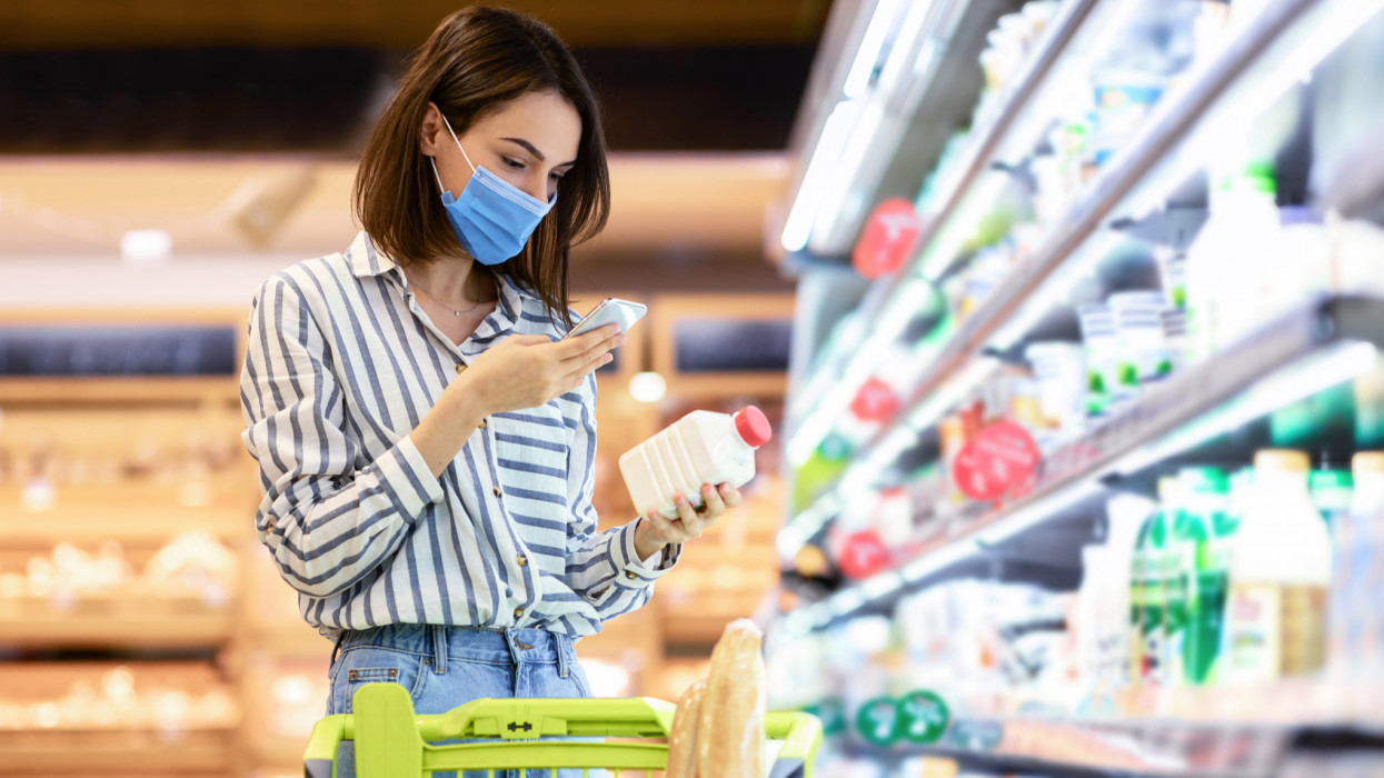 Young woman in disposable face mask taking dairy products from shelf in the supermarket, holding bottle and smartphone, scanning bar code on product through mobile phone, walking with trolley cart