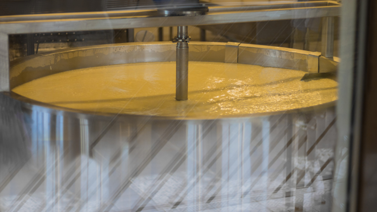 Technology of making cheese from milk on cheese factory, shot through glass of aceptic room, cooking cheese in huge metal tub. Horizontal. Flecks on glass.