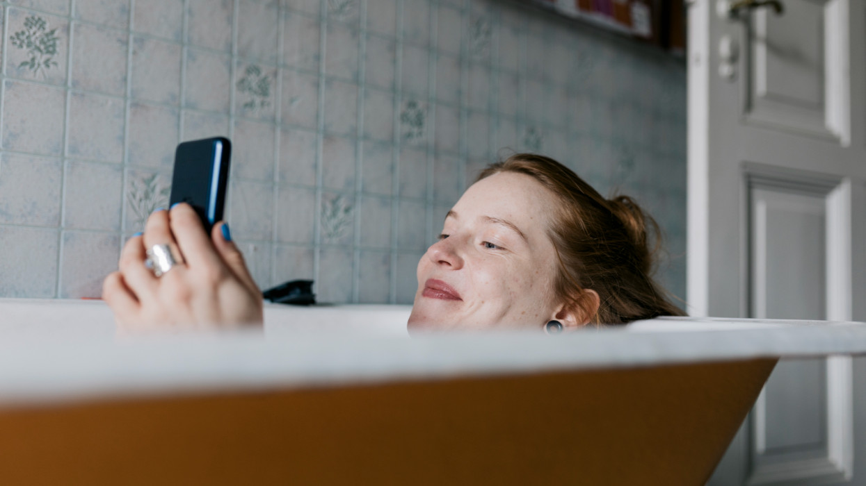 A woman taking a bath and smiling while messaging someone she met on a dating app using her smartphone.