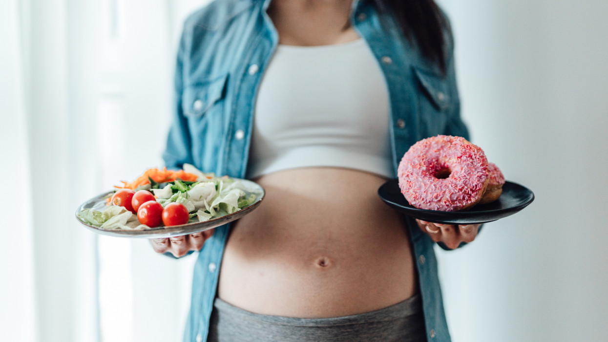 Midsection of pregnant woman choosing between vegetable saiad and doughnuts. Eating well in pregnancy. Food craving during pregnancy concept.