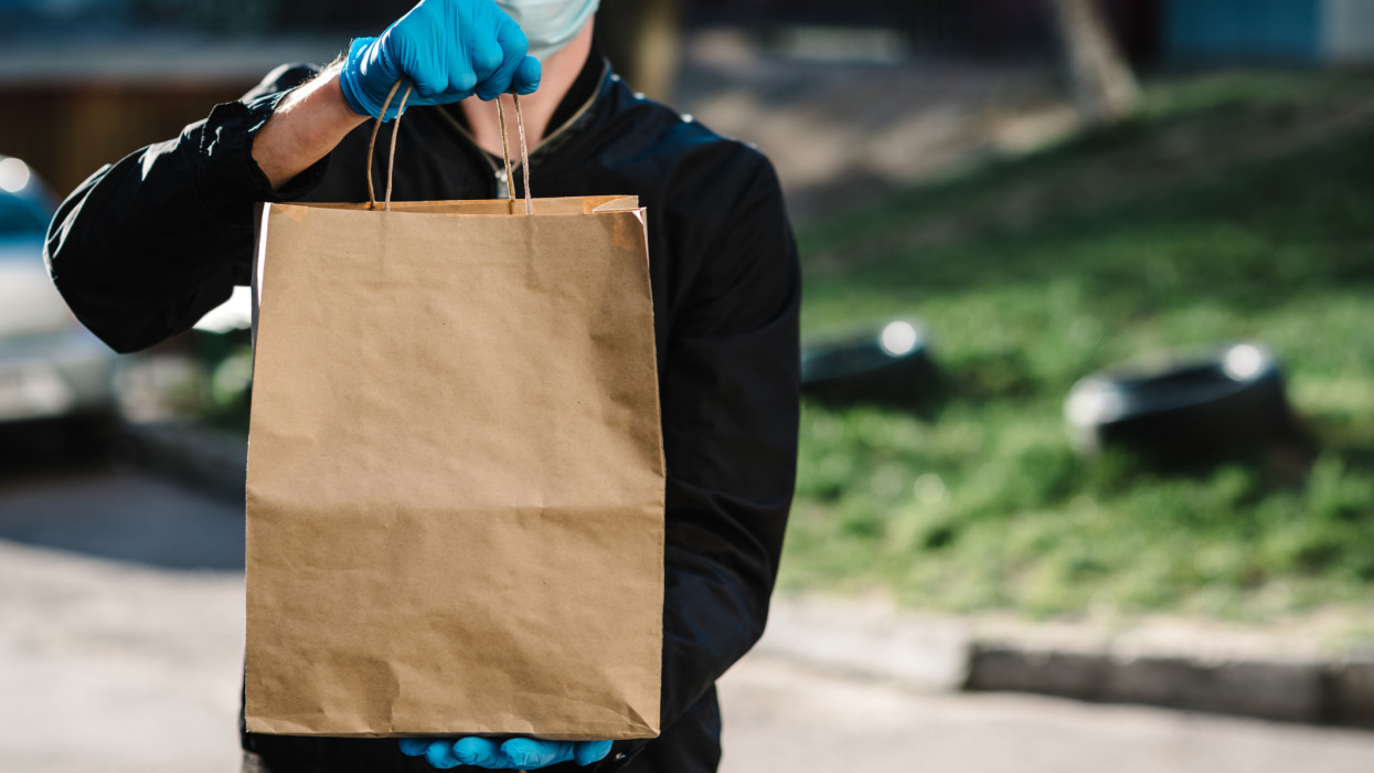 Courier in protective mask, medical gloves delivers takeaway food.  Employee hold cardboard package. Place for text. Delivery service under quarantine, 2019-ncov, pandemic coronavirus, covid-19.