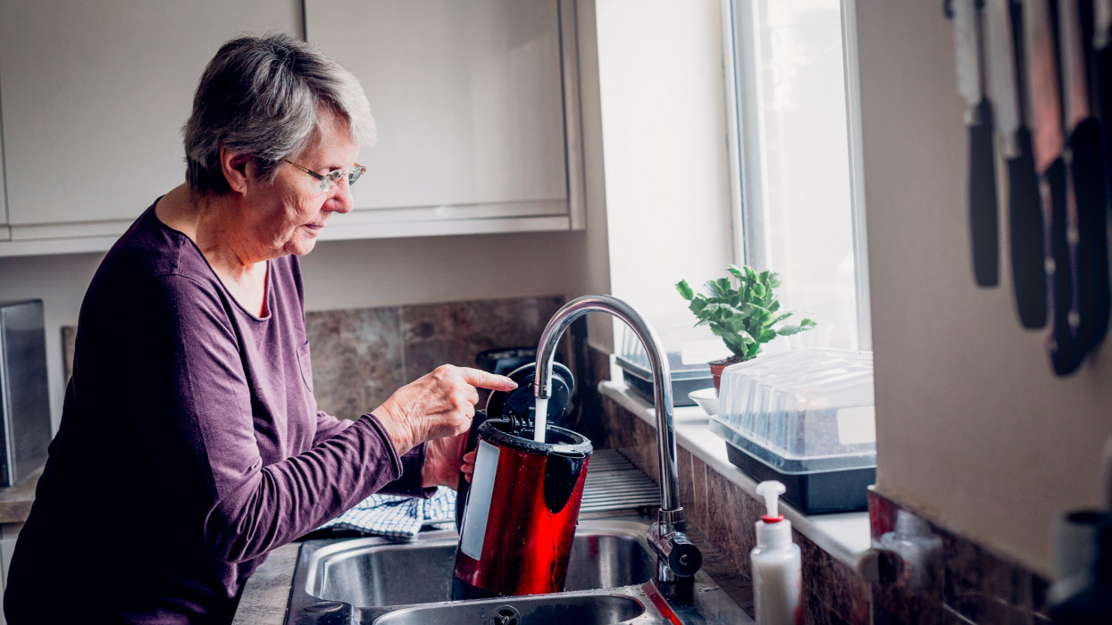 Elderly woman filling up a kettle while standing at her kitchen sink.