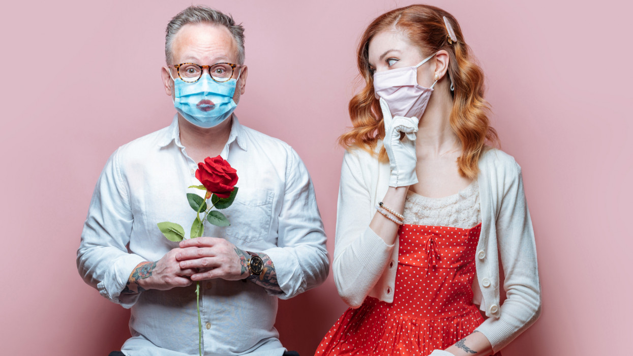 Studio photograph of Valentines Day - couple in Covid-19 quarantine with PPE masks. On pink background. Man holding red roses, and lipstick kiss on PPE mask. Oman with long red hair, wearing red dress and white sweater.