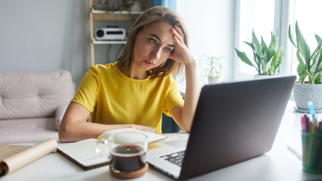 A 35-year-old woman in a bright yellow jacket is sitting in front of a laptop in emotional tension. The concept of working from home, freelance