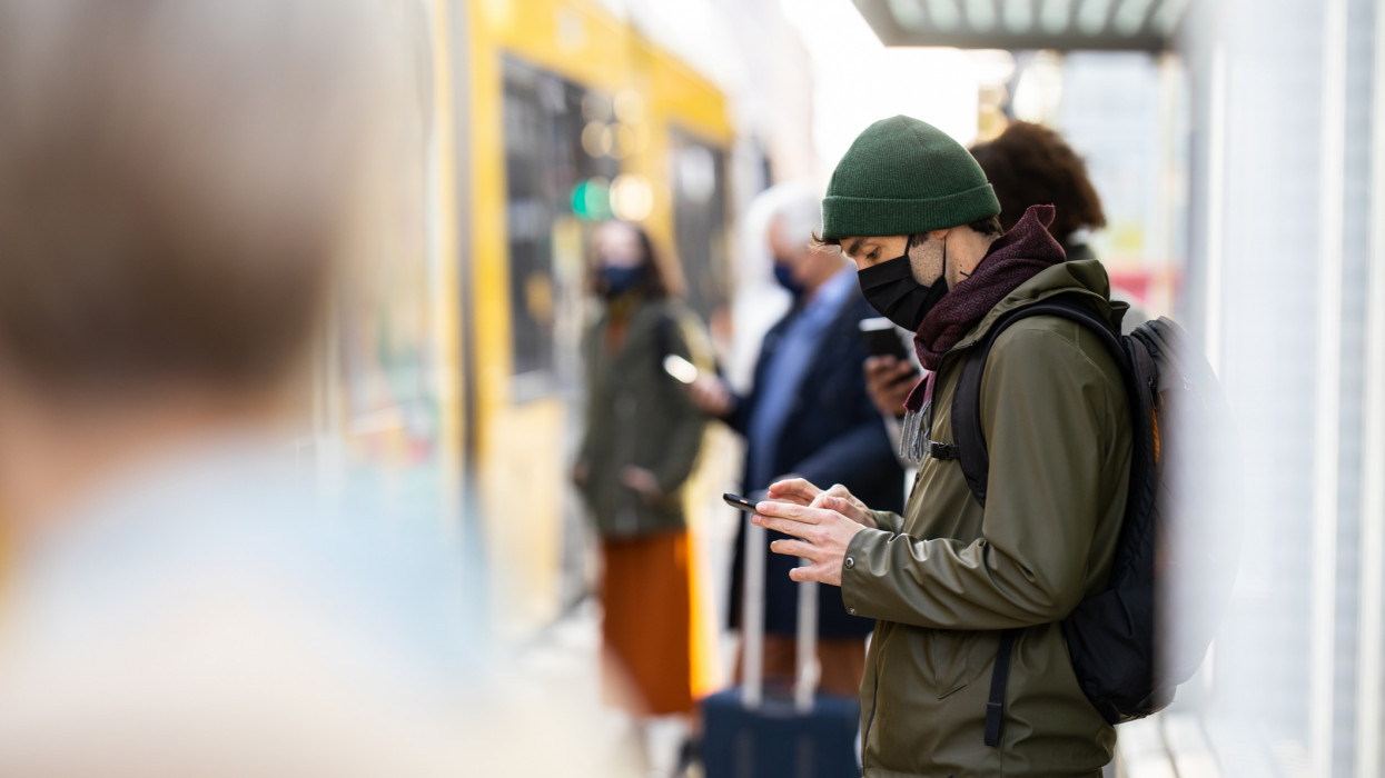 Young man wearing face mask standing at the crowded tram station. People wearing protective face masks standing at city transport station during coronavirus pandemic.