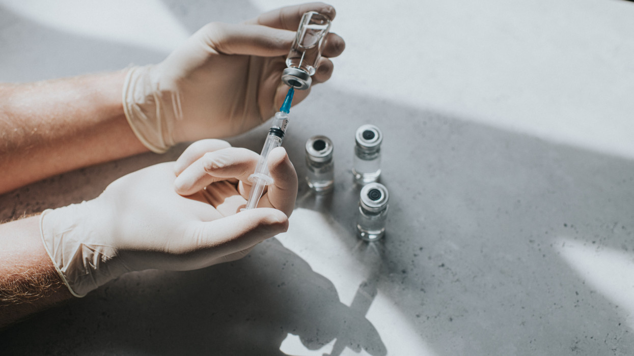 A hand wearing latex gloves hold a phial of clear liquid and a syringe. The medical professional uses the syringe to extract the medicine from the glass bottle. Conceptual with space for copy.