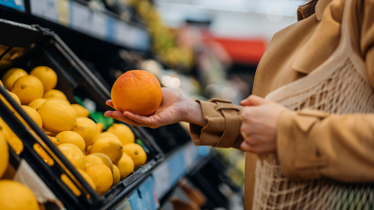 Close up shot of woman shopping for fresh organic fruits and vegetables in supermarket. Putting an orange into eco cotton mesh bag. Zero waste concept.