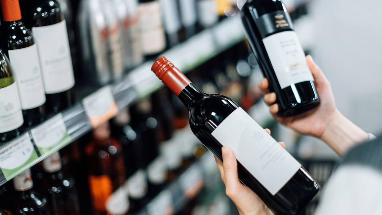 Over the shoulder view of woman walking through liquor aisle and choosing bottles of red wine from the shelf in a supermarket
