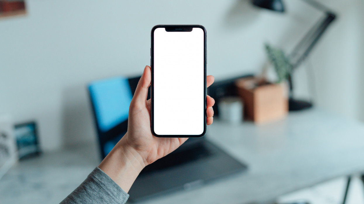 Mockup image of woman holding smartphone with blank white screen at home. Cropped shot of female hand holding phone against home office setup (work desk, laptop and table lamp...) in the study room. Smart home control system.