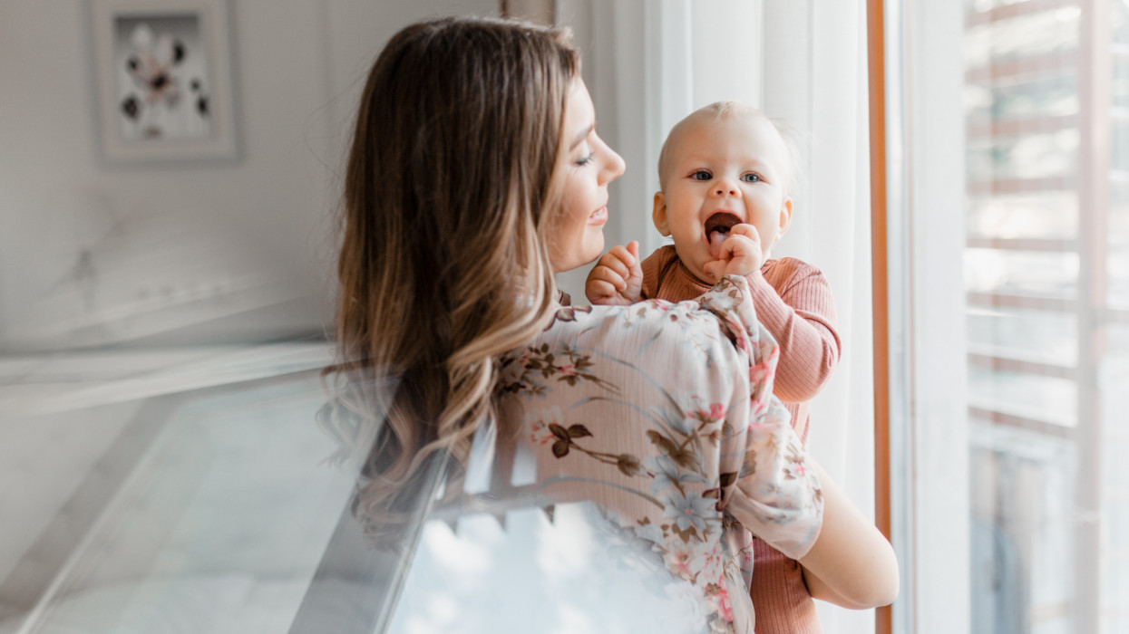 Young woman mom with baby girl on hands near window at home