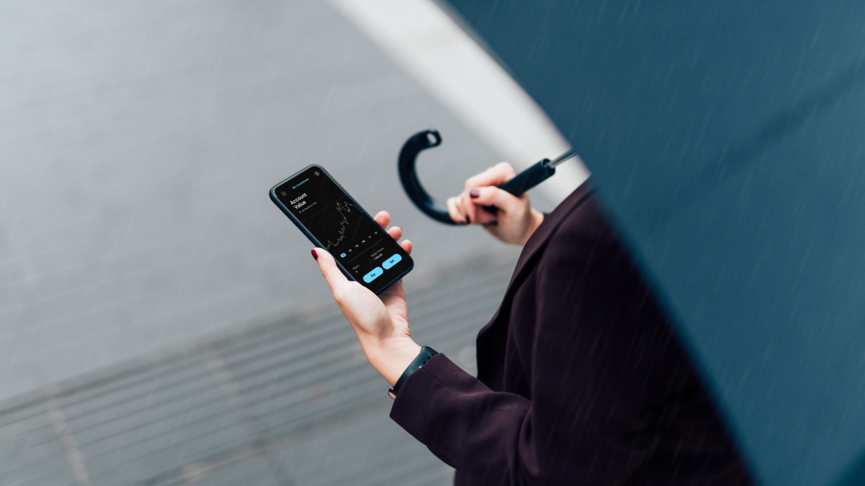 High angle view of a businesswoman using mobile app checking financial data on smartphone, holding an umbrella, walking on the city street in the rain. Insurance protection concept. Business on the go concept.