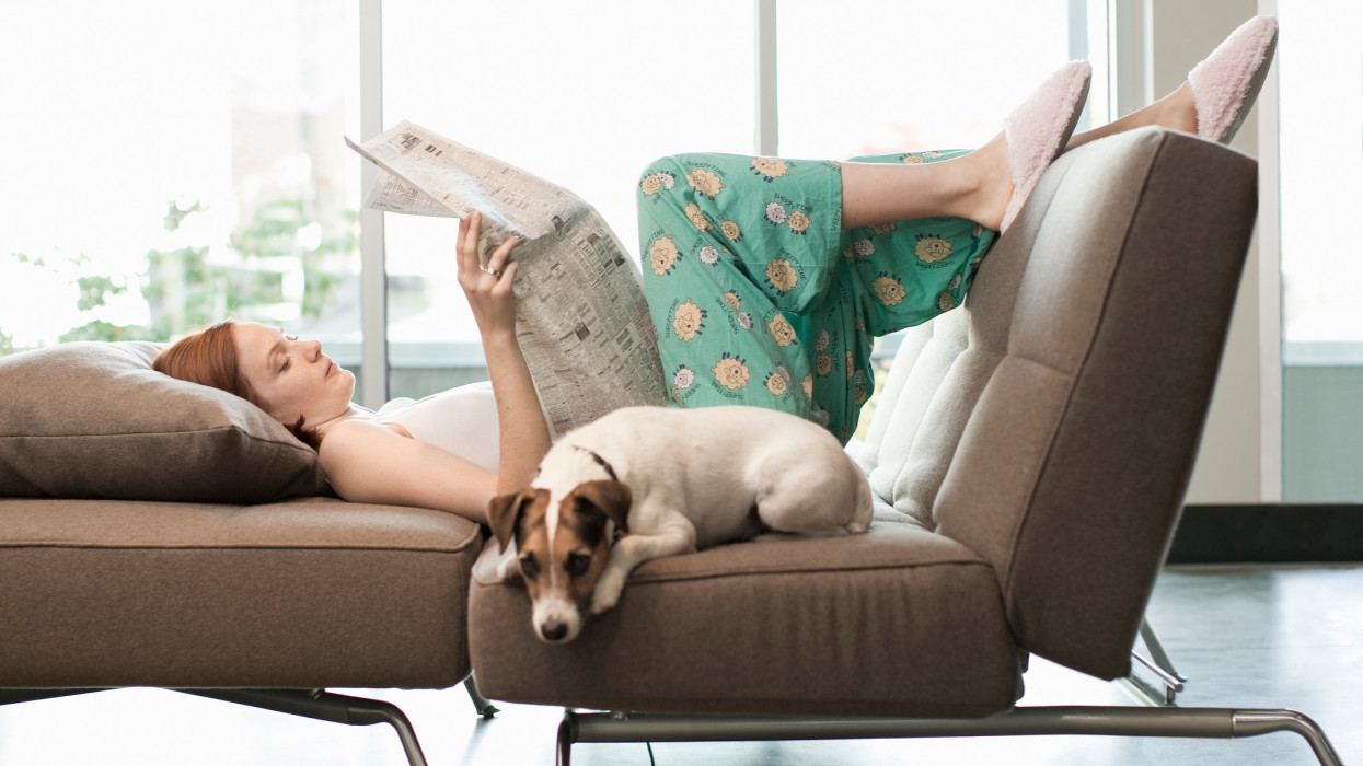 25yr. Cauc woman in pajama bottoms and camisole, laying on couch as she reads newspaper, Jack Russell terrier laying next to her