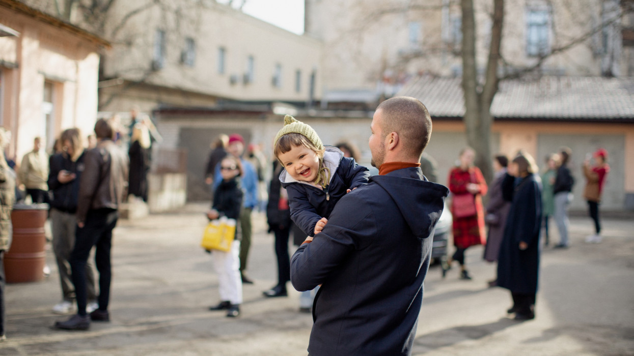 Young man in the blue jacket turned his back to the camera. He holds cute little girl. Girl in the knit hat looks away and laughs cheerfully. People, trees and buildings are on the background.