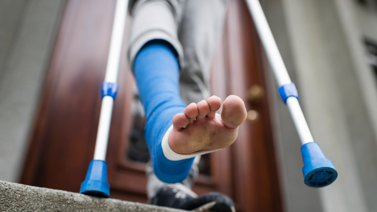 A young adult with crutches and his foot bandaged in plaster is descending the steps of a stairway outside the front door of his apartment. Barefoot toes are sticking out of bandage.