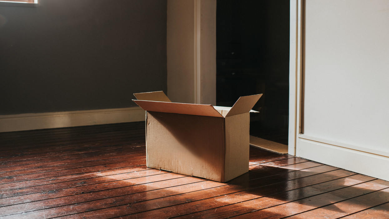 A single, plain, cardboard box sits on a dark wooden floor in a domestic room. Sun shines through the window creating flares and shadows. Room is empty and sparse. Wall provides a space for copy.