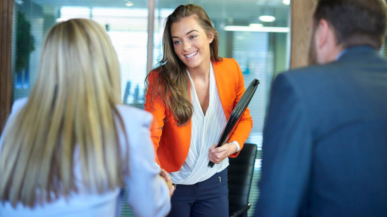 a young graduate walks into an interview room full of confidence and positivity energy . She is holding her cv and smiling at the interview panel before her. She is wearing blue trousers with an orange suit jacket , as she opens the door and strides in and shakes hands with a woman on the panel . in the foreground we can see the back of two of the panel , a man and a woman.