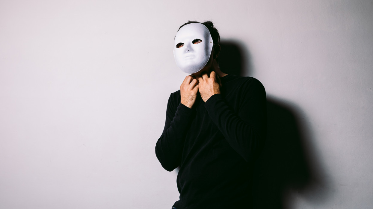 Male dressed in black and wearing white mask over white wall on Halloween holding hands around neck