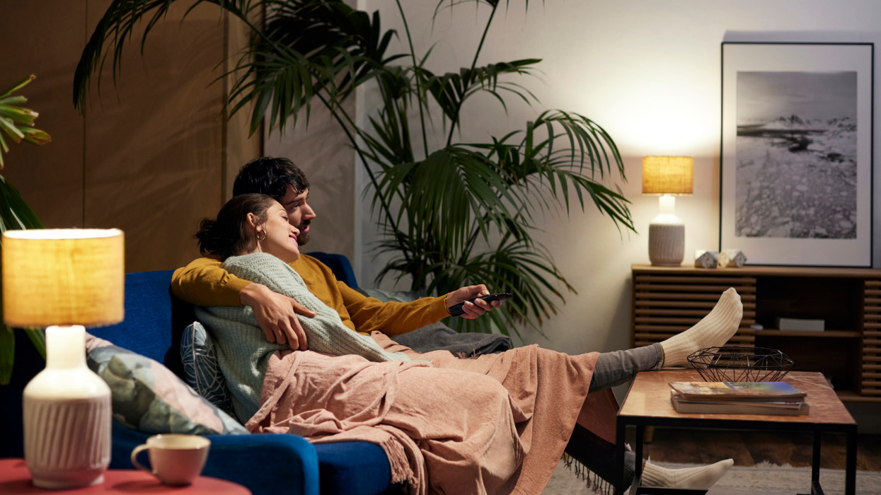 Relaxed couple sitting on sofa in living room. Girlfriend and boyfriend are watching TV at home. They are spending leisure time together.